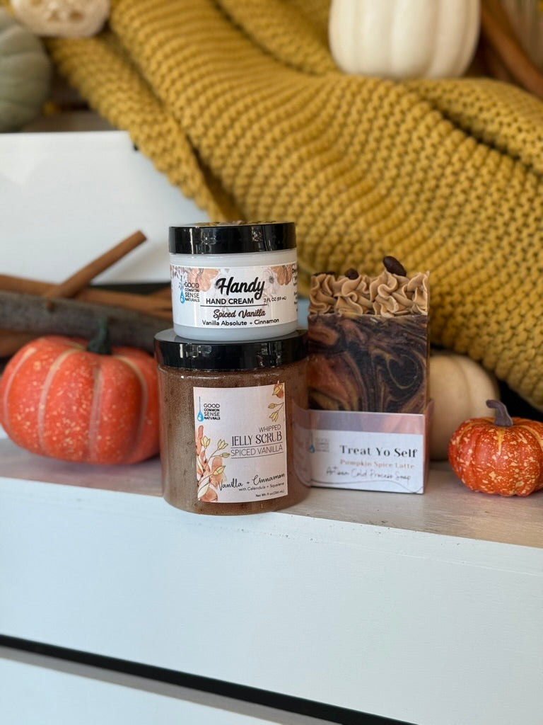 A fall-themed gift set from Handy Candles and The Fly Shop, featuring a jar of Grand Olfact hand cream, a jar of pumpkin spice cinnamon candle, a bag of marbled soap, and a bag of treat yo self pumpkin spice latte soap, styled with pumpkins and a yellow knit blanket.