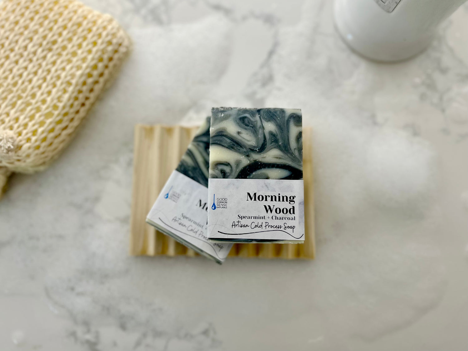 Two bars of Morning Wood Cold Process Bar Soap, featuring spearmint and charcoal, rest on a wooden dish amid suds—ideal for acne-prone skin. A textured washcloth and white bottle sit nearby, all arranged on a white surface.
