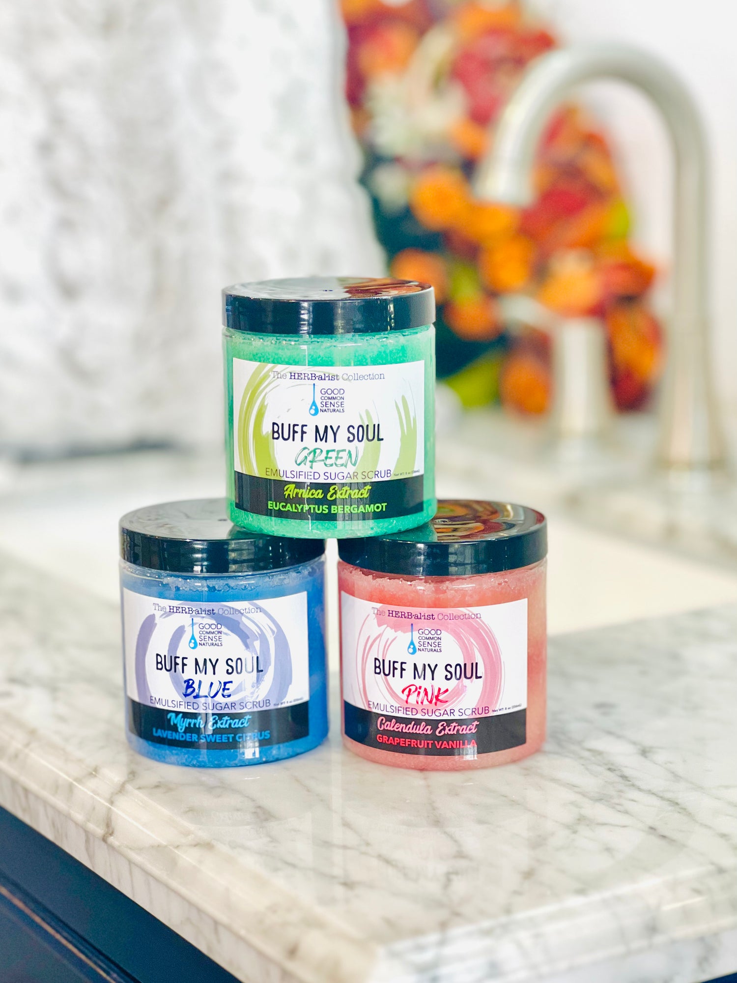 Three jars of Buff My Soul sugar scrub in blue, green, and red colors, placed on a marble surface.