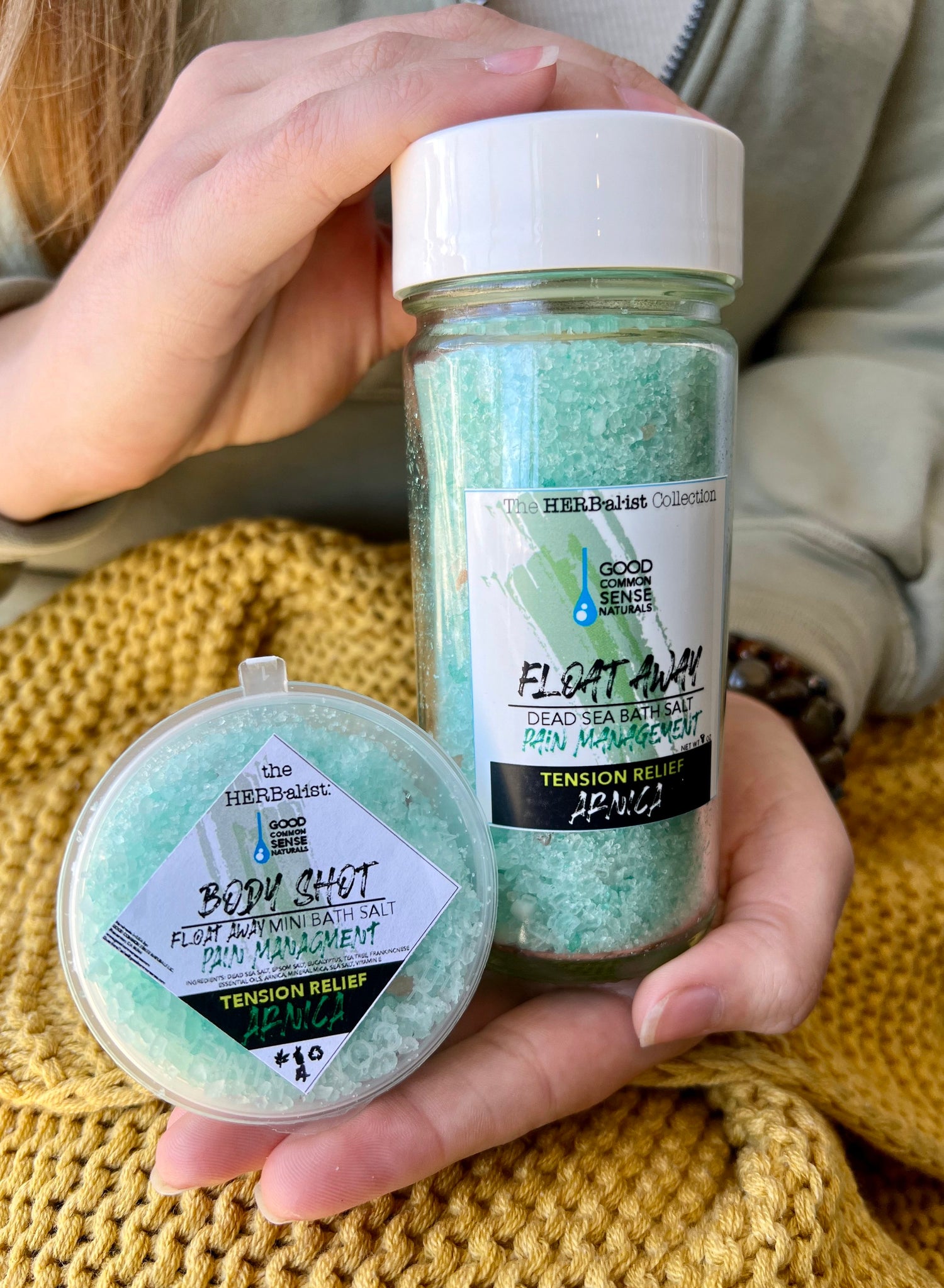 Two containers of bath salts with eucalyptus and tension relief properties, held in a person's hand. One larger container is labeled 'Float Away' and the other is a smaller shaker bottle
