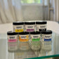 A collection of seven small, 2 oz jars of Handy brand lotion in various colors, each with a different scent.