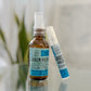 Two Grain Rain Hand Sanitizer bottles, one regular and one travel spray, stand on a white surface. Their blue labels with black text pair with a softly blurred background of green leaves, creating a fresh atmosphere.