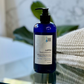 A blue pump bottle of shampoo labeled 'Roots To Recovery - Tea Tree & Rosemary Shampoo, 16oz'. The bottle is placed on a reflective surface with a textured background.