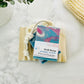 The Well Hung bar soap, with marble swirls of blue, pink, and white, sits on a wooden dish with a rope attached. Nearby are a plant and exfoliating pouch, showcasing this artisan eucalyptus lavender soap on a rope.