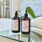 Two bottles of hair care products, one labeled as 'Shampoo' and the other as 'Conditioner', with pump dispensers, placed on a surface with a plant and a blurred background.