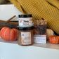 A fall-themed gift set from Handy Candles and The Fly Shop, featuring a jar of Grand Olfact hand cream, a jar of pumpkin spice cinnamon candle, a bag of marbled soap, and a bag of treat yo self pumpkin spice latte soap, styled with pumpkins and a yellow knit blanket.