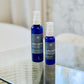 Two blue bottles of Good Light Evening Moisturizer with spray nozzles, one larger than the other, placed on a glass surface with a blurred background.