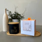 A Butter Massage Candle Simply joyfull, alongside a custom-made glass jar with a spout, on a wooden tray, with a plant in the background.