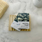 Two bars of Morning Wood Cold Process Bar Soap, featuring spearmint and charcoal, rest on a wooden dish amid suds—ideal for acne-prone skin. A textured washcloth and white bottle sit nearby, all arranged on a white surface.