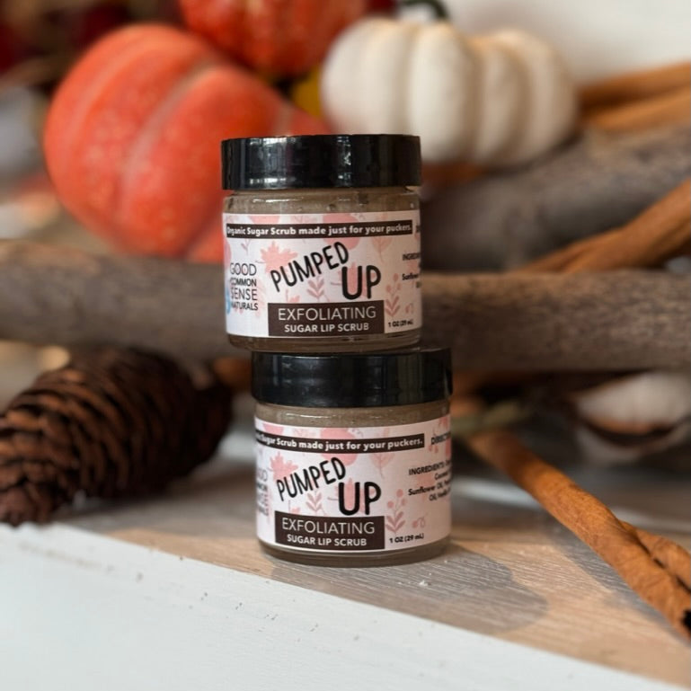 Two jars of Good Common Sense Naturals Pumped Up Exfoliating Sugar Lip Scrub are stacked on a white display ledge, surrounded by cozy fall décor including pumpkins, cinnamon sticks, and a pinecone. The warm, softly blurred background creates an inviting autumn atmosphere that highlights the handcrafted seasonal lip scrub.