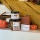 The Glow Up Fall Into Spice gift set featuring Fall Into Spice Handy Hand Cream, Pumpkin Queen Whipped Soap Scrub, and Oh Gourd Yass! artisan soap displayed on a white shelf with miniature pumpkins, cinnamon sticks, cotton stems, and a cozy mustard knit blanket in the background. The warm autumn display creates an inviting seasonal spa aesthetic.