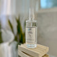 A clear spray bottle with a white label containing an aromatherapy blend, placed on a wooden stand on top of a glass table.