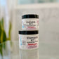 Two jars of Disappearing Act 1 Skin Toning Cream with labels indicating the product name and ingredients such as cocoa butter, shea butter, and essential oils.