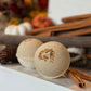 Two round bath bombs with a warm beige color and a brown spice pattern on top, displayed on a white wooden surface with cinnamon sticks and autumn decorations in the background.