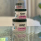 Three containers of Handy brand hand cream in different sizes placed on a reflective surface with a plant in the background.