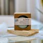 A solid shampoo bar packaged in a cardboard box with a clear window, placed on a wooden soap dish 