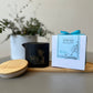 A Butter Massage Candle - Hippie's Brew is displayed with its packaging and a wooden dish, accompanied by a potted plant and a decorative blue ribbon.