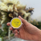 Hand holding a small round container with a yellow label against a blurred natural background