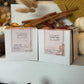 Two white boxed Butter Massage Candles sit side by side on a white display ledge in a cozy fall retail window. The left box is tied with a gold ribbon and labeled Vanilla Chai, while the right box is tied with a deep burgundy ribbon and labeled Fall Into Spice. Behind them, cinnamon sticks, dried wheat stems, knit fabric, and white heirloom pumpkins create a warm autumn backdrop with soft golden bokeh. A pinecone rests in the foreground, adding seasonal texture. The shallow depth of field keeps the products