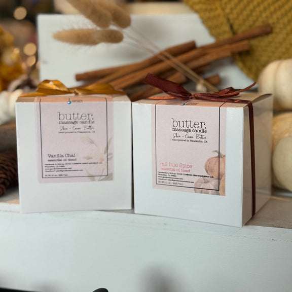 Two white boxed Butter Massage Candles sit side by side on a white display ledge in a cozy fall retail window. The left box is tied with a gold ribbon and labeled Vanilla Chai, while the right box is tied with a deep burgundy ribbon and labeled Fall Into Spice. Behind them, cinnamon sticks, dried wheat stems, knit fabric, and white heirloom pumpkins create a warm autumn backdrop with soft golden bokeh. A pinecone rests in the foreground, adding seasonal texture. The shallow depth of field keeps the products
