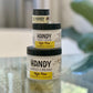 Three Handy Cream - High Pine containers in various sizes are stacked on glass. The label features Sandalwood + Lavender. A blurred green plant in the background accents this nourishing hand cream display.