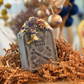 Life After Death bath bomb shaped like a weathered headstone, blooming with rose petals and blue flowers as it fizzes, Good Common Sense Naturals