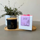 A black candle with a purple label on top, placed on a wooden dish with a floral decoration in the background.