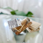 A cleansing bundle consisting of a sage bundle, selenite wand, and palo Santo placed on a glass surface, with a label in the center.