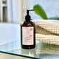 A bottle of liquid conditioner with a pump dispenser, placed on a glass surface, with a white towel and a plant in the background.