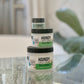 Three containers of Handy Cream - Sauna Rescue, enriched with eucalyptus and bergamot oils, are stacked on a glass table. A blurred plant and soft lighting in the background set a refreshing, revitalizing scene.