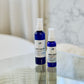 Two blue bottles of Moisturizer with spray nozzles, one larger than the other, placed on a glass surface with a blurred background.