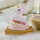 A pegasus-shaped bath truffle with a pink and white color scheme, placed on a wooden surface with bubbles around it, suggesting it is a bath toy designed to create bubbles.