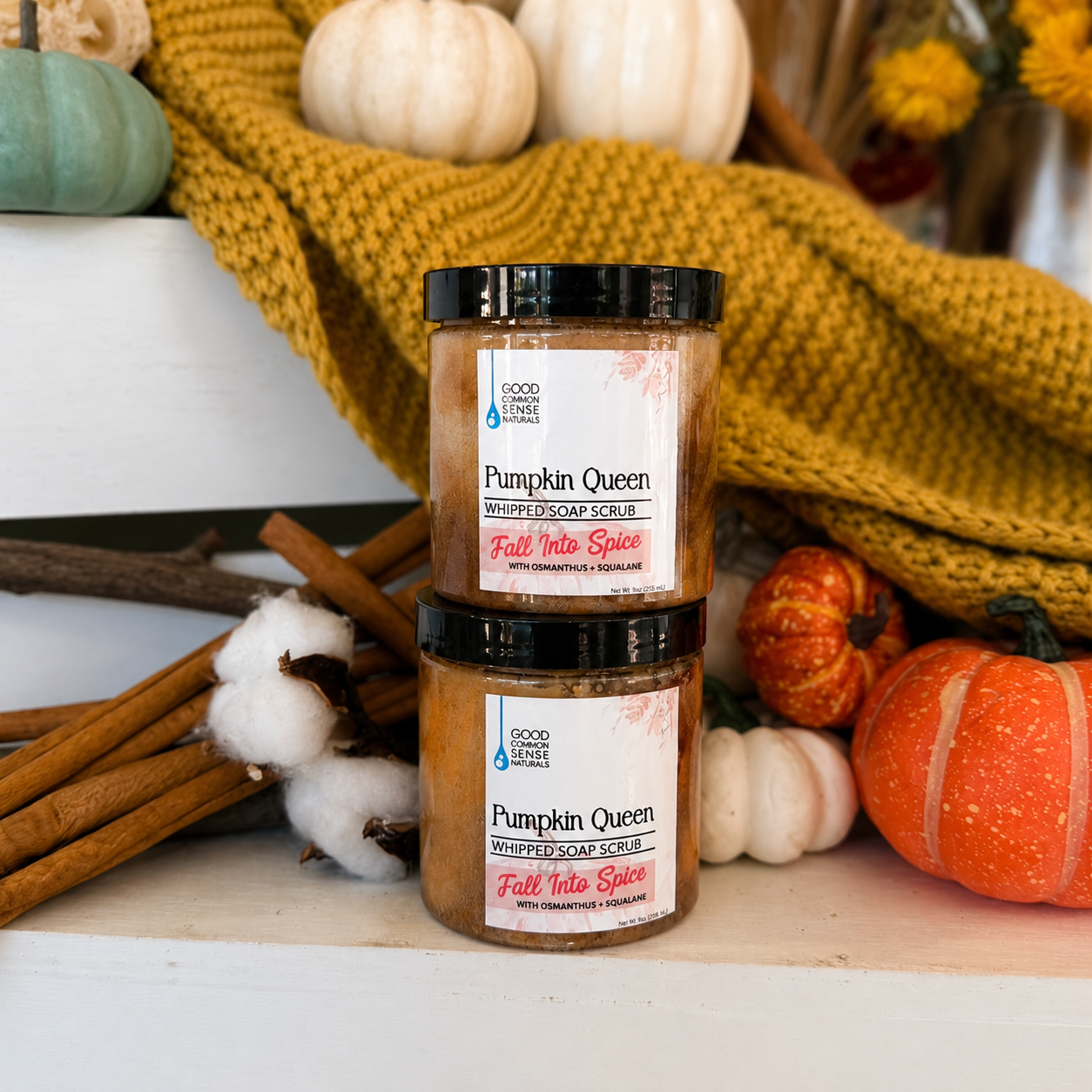 Two jars of Good For You Soap Co. Pumpkin Queen Fall Vibes soap scrub stacked on a white surface, surrounded by autumn props including pumpkins, a yellow knit blanket, cinnamon sticks, and cotton bolls.