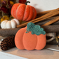 A pumpkin-shaped orange bath bomb with a textured dark green stem and leaf detail sits on a white wooden shelf, styled among rustic fall décor including cinnamon sticks, pinecones, cotton bolls, and decorative pumpkins. The softly blurred background features warm autumn florals and seasonal accents, creating a cozy harvest-inspired atmosphere that highlights the bath bomb as the focal point.