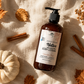 A warm, autumn-themed flat lay featuring an amber pump bottle of Good Common Sense Naturals Native Skin Soothing Body Lotion, Vanilla Chai Limited Edition, resting on textured cream-colored fabric. The lotion bottle is surrounded by a white pumpkin, scattered orange maple leaves, cinnamon sticks, a dried botanical sprig, and soft golden sunlight casting dappled shadows across the scene. The cozy seasonal styling emphasizes fall warmth while keeping the lotion bottle as the central focus.