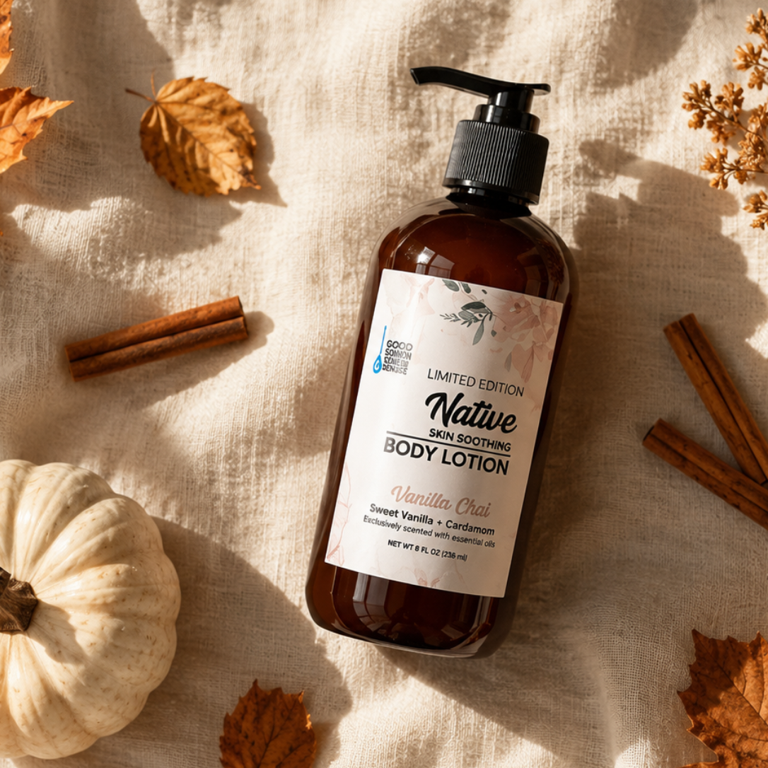 A warm, autumn-themed flat lay featuring an amber pump bottle of Good Common Sense Naturals Native Skin Soothing Body Lotion, Vanilla Chai Limited Edition, resting on textured cream-colored fabric. The lotion bottle is surrounded by a white pumpkin, scattered orange maple leaves, cinnamon sticks, a dried botanical sprig, and soft golden sunlight casting dappled shadows across the scene. The cozy seasonal styling emphasizes fall warmth while keeping the lotion bottle as the central focus.