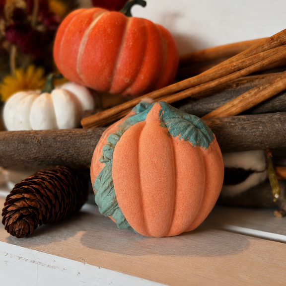 An orange decorative foam pumpkin with a teal-green stem sits on a rustic wooden surface next to a pinecone, with other fall-themed props including cinnamon sticks, a small white pumpkin, and dried flowers in the background.