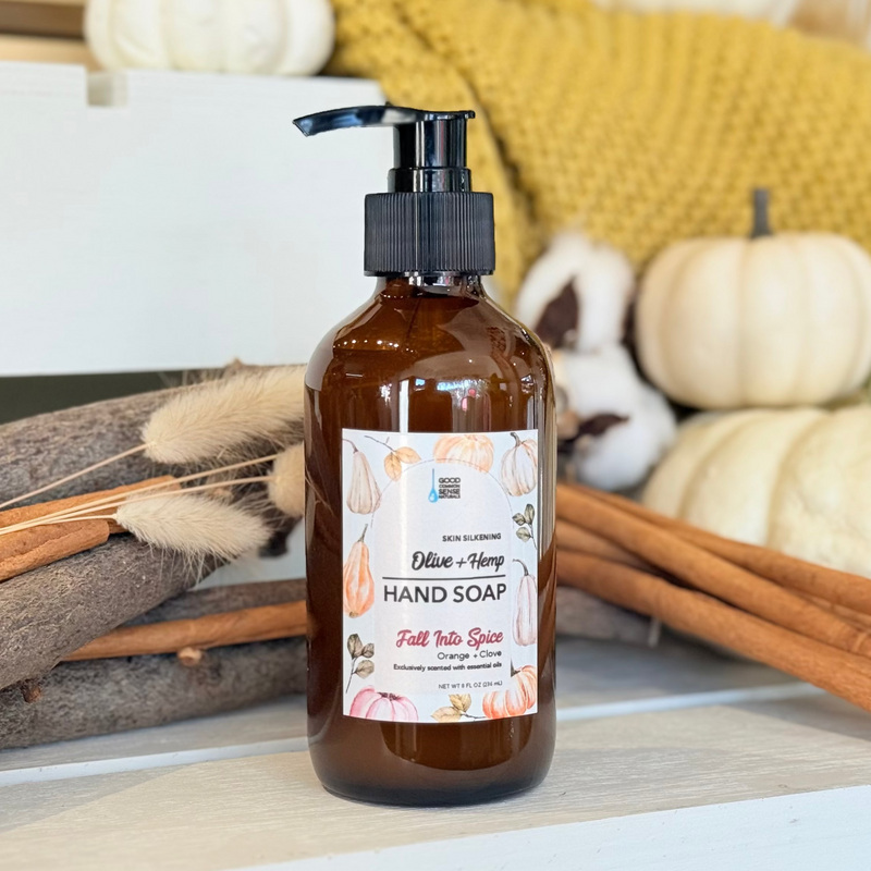 An amber glass pump bottle of Colby & Honey Hand Soap labeled 'Pumpkin Spice' sits on a white surface surrounded by fall-themed props including cinnamon sticks, dried grass, cotton bolls, and small pumpkins.