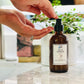 A person's hand holding a bottle of liquid soap with a pump lid, over a marble surface with a plant in the background.