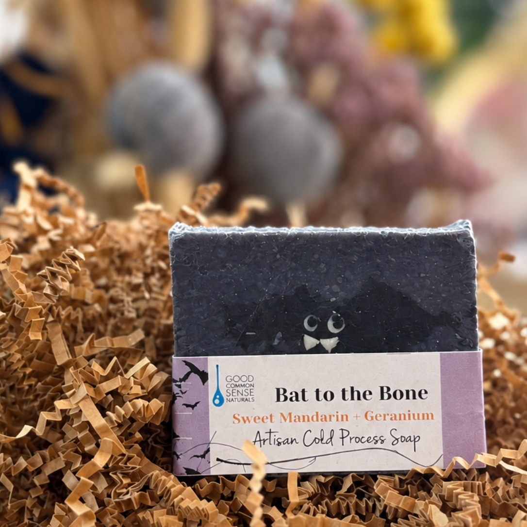 A rectangular bar of soap labeled 'Bat to the Bone' sits centered in brown shredded paper filler on a wooden tray, with a softly blurred decorative background.