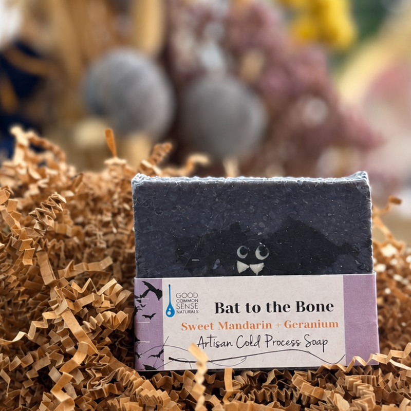 A rectangular bar of soap labeled 'Bat to the Bone' sits centered in brown shredded paper filler on a wooden tray, with a softly blurred decorative background.