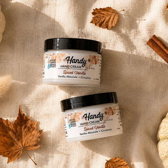 Two jars of Handy Hand Cream in Sweet Vanilla scent, arranged in a styled flat-lay with pumpkins, cinnamon sticks, and autumn leaves on a textured cream fabric background.