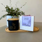 A Butter Massage Candle labled Lavender honey alongside a custom-made glass jar with a spout, on a wooden tray, with a plant in the background.