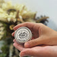 Hand holding a small metal container with a decorative label against a blurred natural background