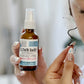 A bottle of 'Blown Away - Blow Out' spray for frizz control, held in a hand with a manicure. The bottle is clear and has a label with the product name and a brief description.