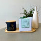 A Butter Massage Candle with Sauna Rescue Eucalyptus & Lemon scent, alongside a custom-made glass jar with a spout, on a wooden tray, with a plant in the background.