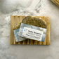 A bar of handmade soap with 'Salty Seamen' text and sea-themed design on the packaging, resting on a wooden soap dish with a marble background.