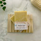 The Sweet Teat Cold Process Bar Soap, made with goat milk and honey, rests on a wooden dish. Crafted from natural ingredients for sensitive skin, it's displayed with a plant, textured cloth, and marble surface.
