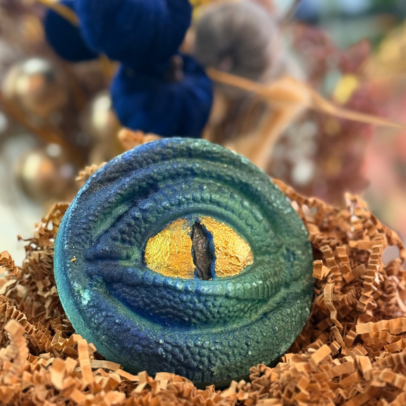 A decorative round bath bomb with a reptile-like eye design, featuring a blue and green textured surface and a gold center, displayed on brown shredded paper with a blurred background of blue and neutral decor.