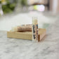 A stick of Vanilla Latte Lip Balm is displayed in a clear tube with a label, placed on a wooden holder on a marble surface.